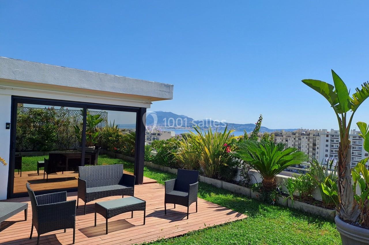 Terrasse avec mobilier d'extérieur, plantes et vue sur une ville côtière sous un ciel bleu dégagé.