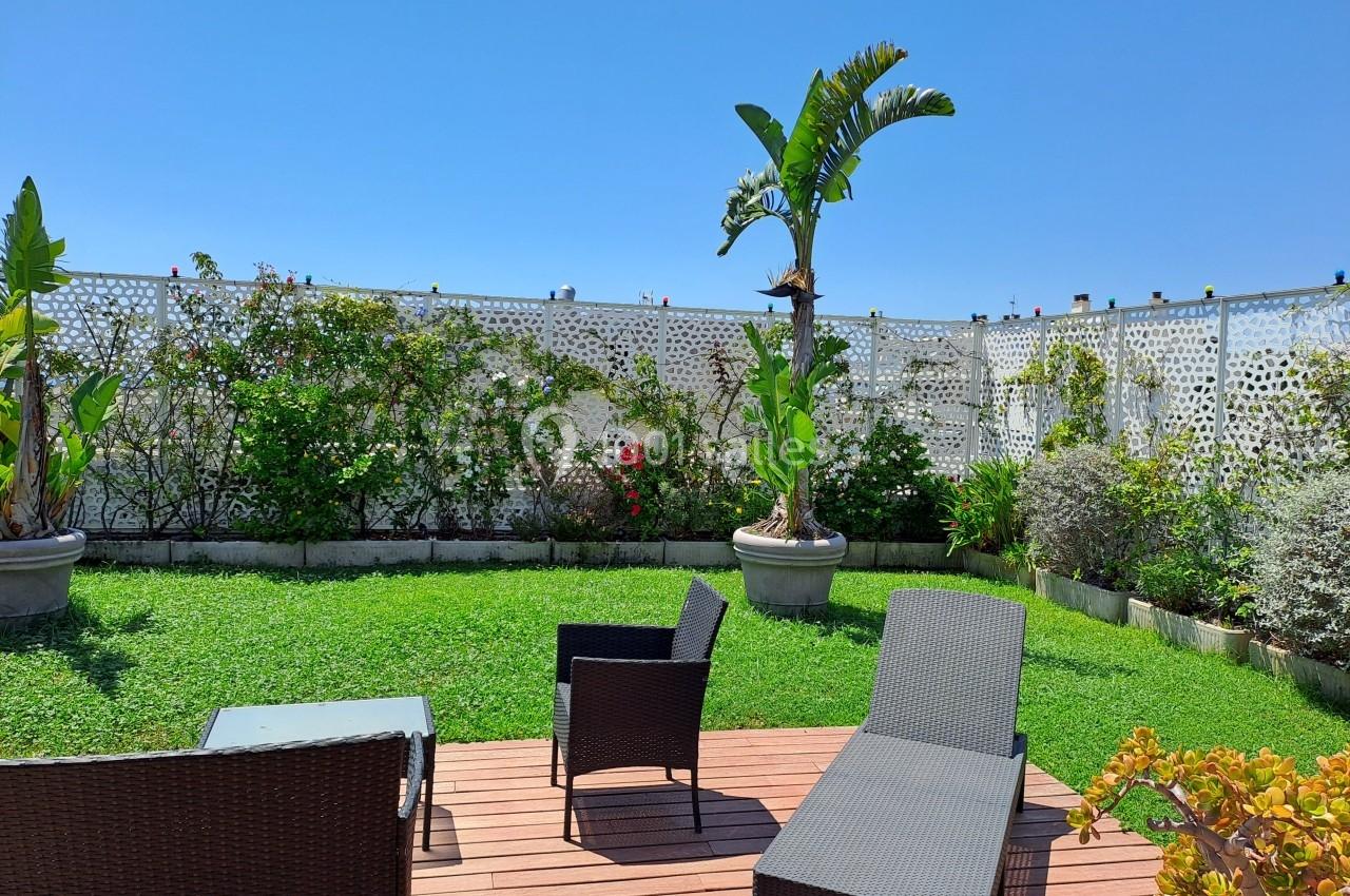 Espace extérieur avec terrasse en bois, chaises en rotin, pelouse, plantes en pot et mur végétalisé sous un ciel bleu.