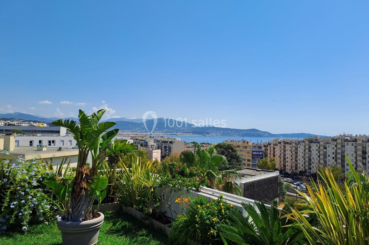 Vue depuis une terrasse avec des plantes en pot, donnant sur des bâtiments, la mer et des montagnes sous un ciel bleu.