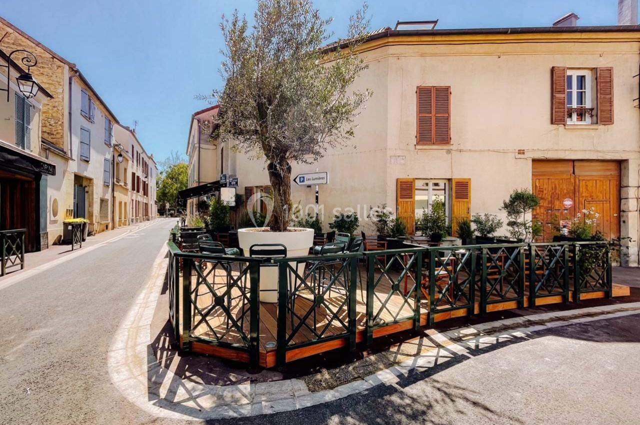 Terrasse en bois avec des tables et chaises, entourée de plantes, située à l'angle d'une rue calme et ensoleillée.