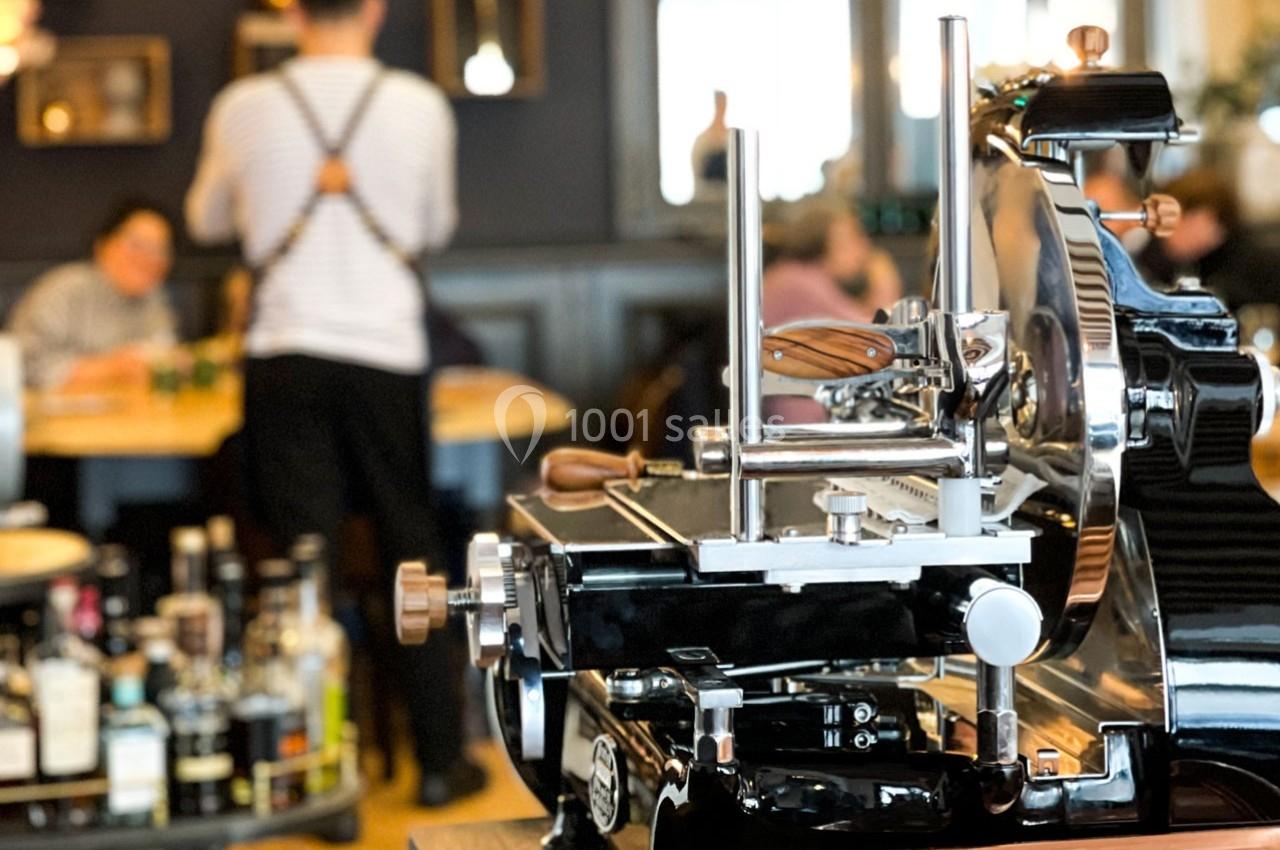 Trancheuse à jambon brillante au premier plan dans un restaurant, avec des clients et un serveur en arrière-plan flou.