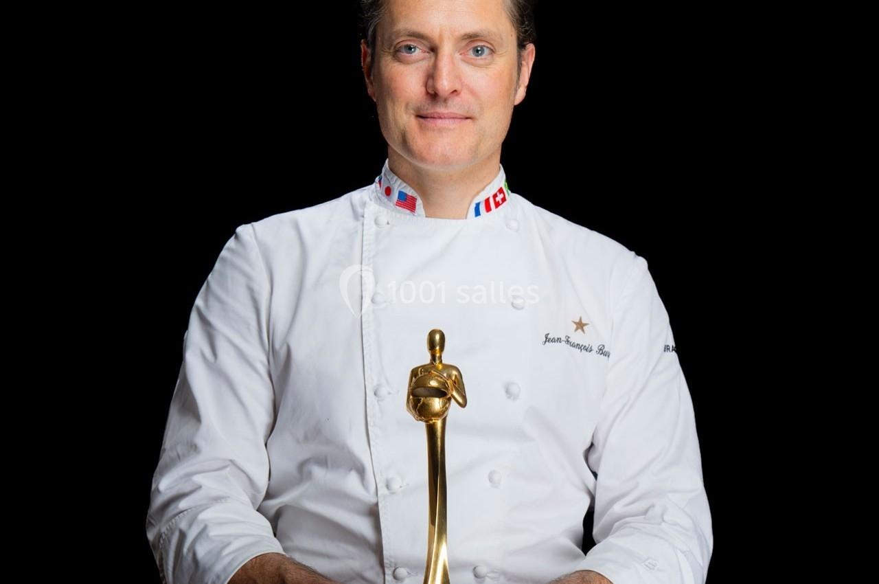 Un chef en veste blanche tient un trophée doré sur un fond noir.