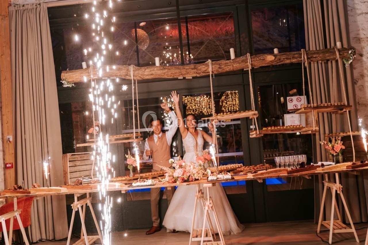 Un couple en tenue de mariage célèbre devant une table décorée de desserts, avec des étincelles en premier plan.
