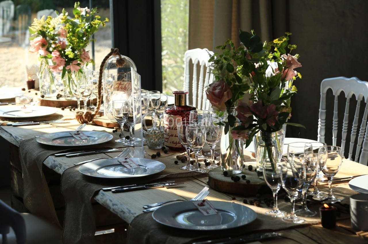 Table en bois décorée pour un repas, avec vaisselle, verres, fleurs et lumière naturelle entrant par une baie vitrée.