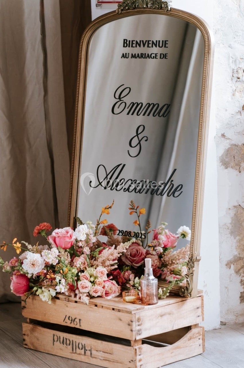 Miroir décoratif avec inscription de bienvenue pour un mariage, entouré de fleurs et posé sur des caisses en bois.