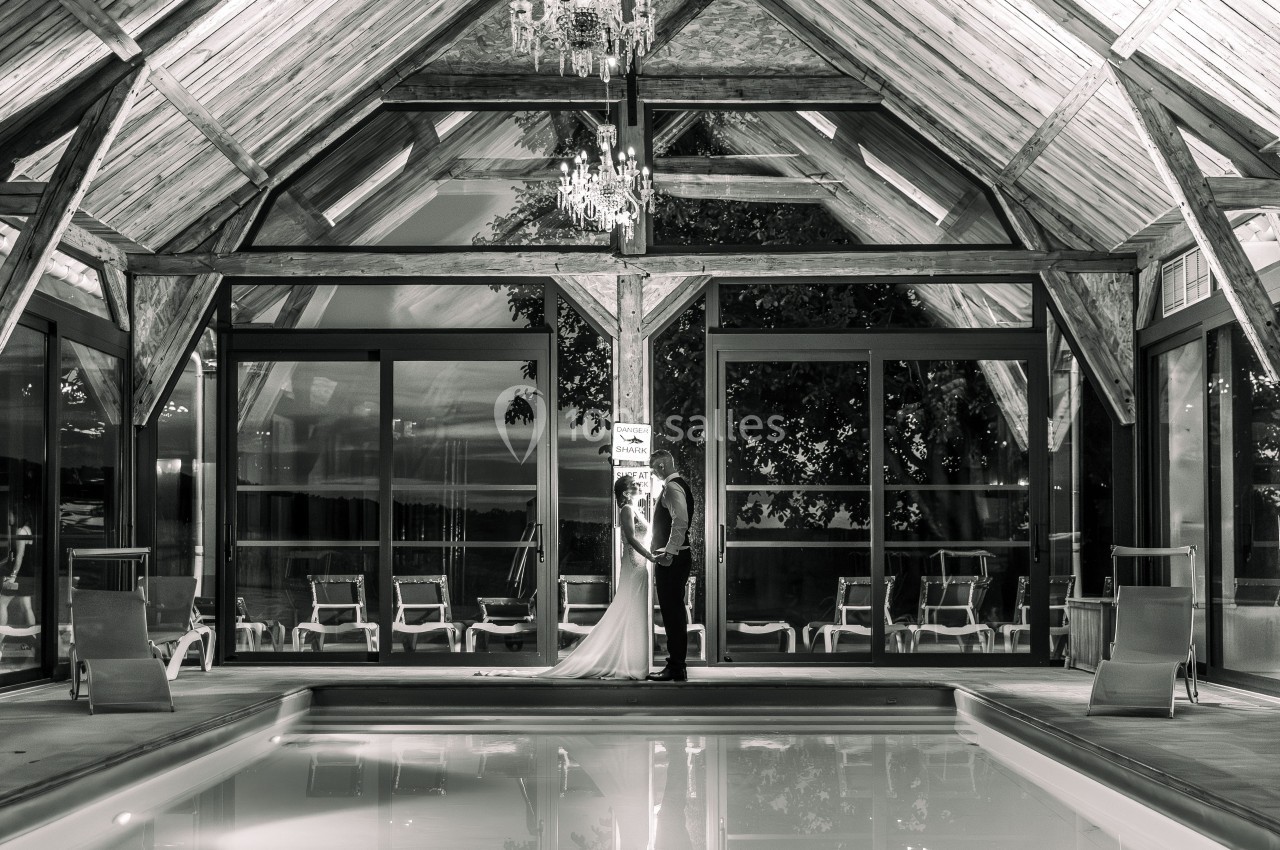 Un couple debout près d'une piscine intérieure, entouré de grandes baies vitrées et d'une structure en bois.