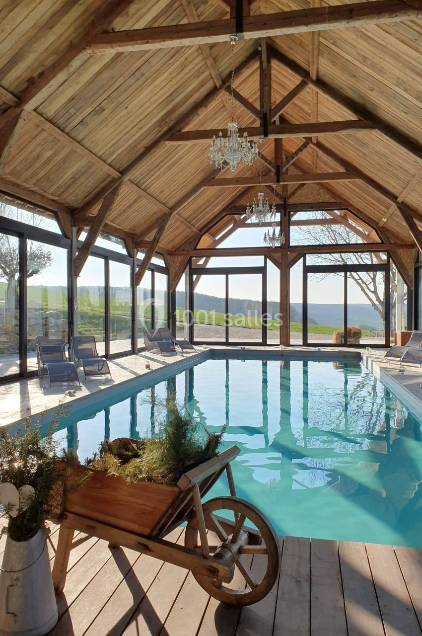 Piscine intérieure lumineuse sous une structure en bois avec de grandes baies vitrées donnant sur un paysage champêtre.