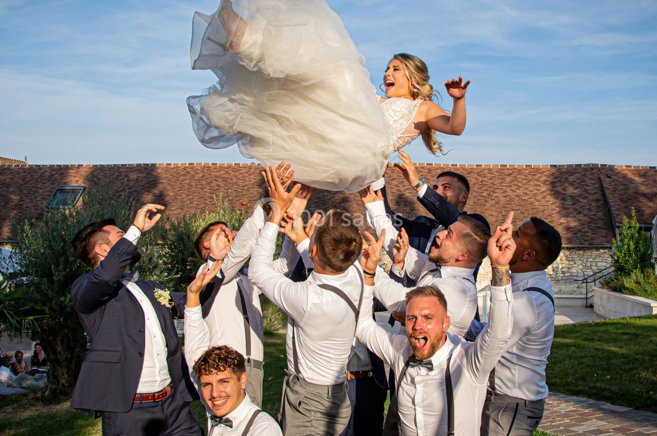 Un groupe d'hommes en tenue de cérémonie soulève une femme en robe blanche dans un jardin par temps ensoleillé.
