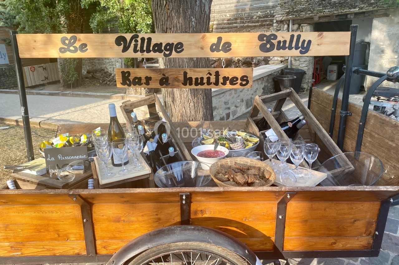 Chariot en bois avec des huîtres, verres, bouteilles de vin et pancarte indiquant ’Le Village de Sully, Bar à huîtres’.