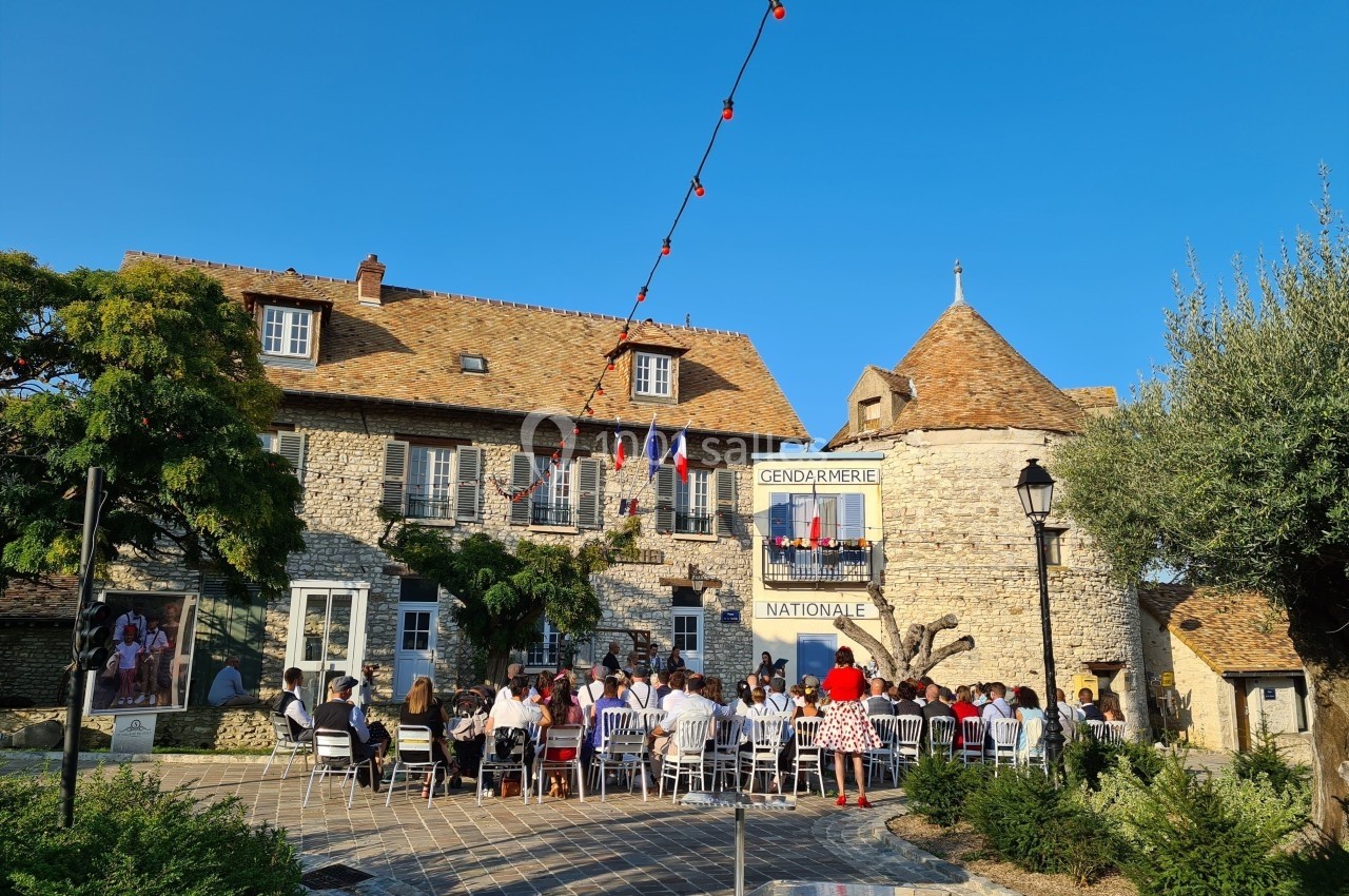 Groupe de personnes assises en extérieur devant un bâtiment en pierre avec des drapeaux et une enseigne ’Gendarmerie…