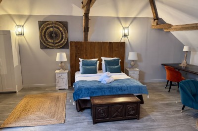 Chambre spacieuse sous combles avec poutres apparentes, lit double, coin salon coloré et grande baie vitrée.