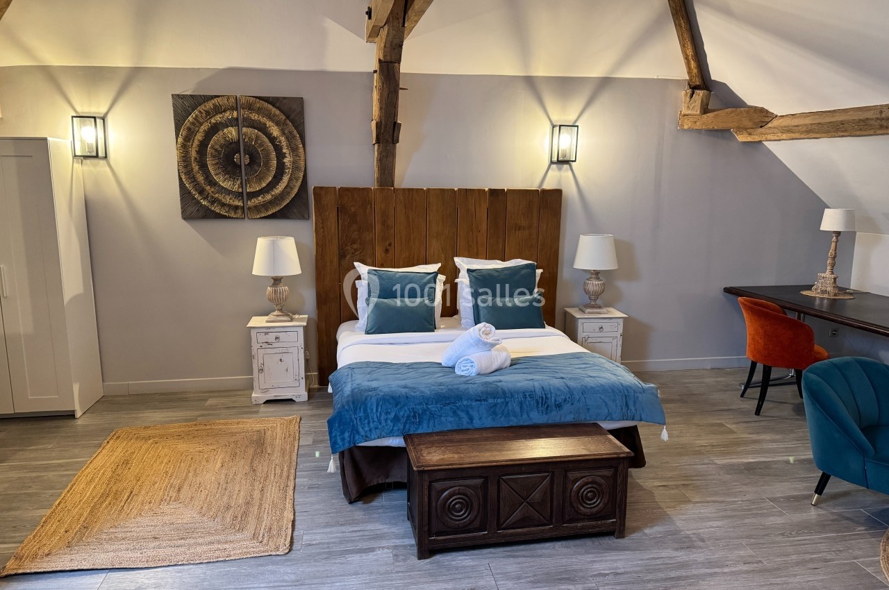 Chambre lumineuse avec lit double, poutres apparentes, mobilier en bois, fauteuils colorés et décoration murale.