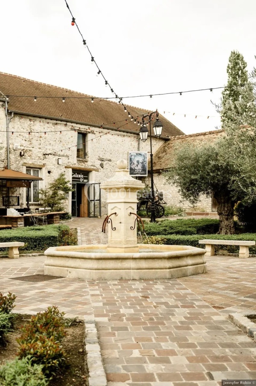 Place pavée avec une fontaine centrale, entourée de bâtiments anciens, de verdure et de guirlandes lumineuses.