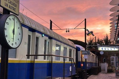 Chambre spacieuse sous combles avec poutres apparentes, lit double, coin salon coloré et grande baie vitrée.