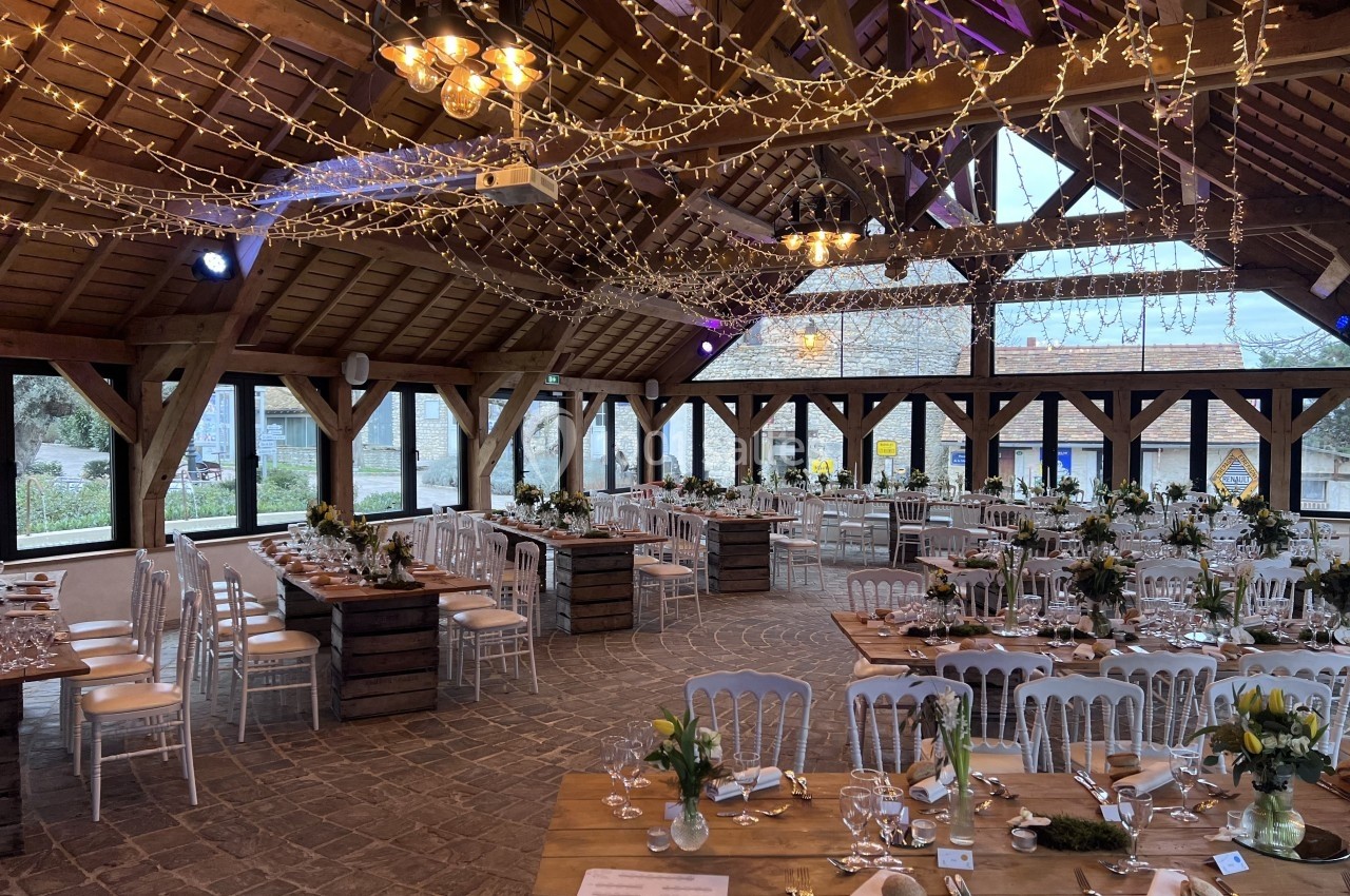 Salle de réception en bois décorée de guirlandes lumineuses, tables dressées avec vaisselle et fleurs.