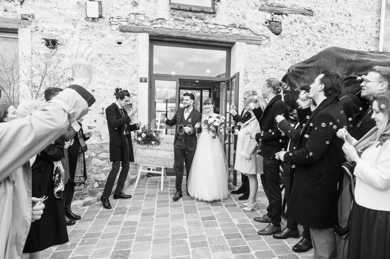 Un couple de mariés sort d'un bâtiment en pierre, entouré d'invités applaudissant sur une allée pavée.