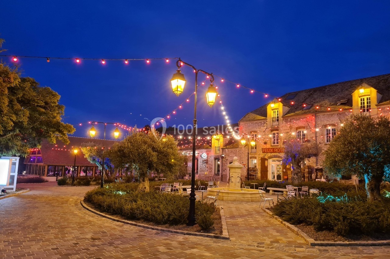 Place pavée illuminée par des guirlandes et lampadaires, entourée de bâtiments en pierre et d'arbres.
