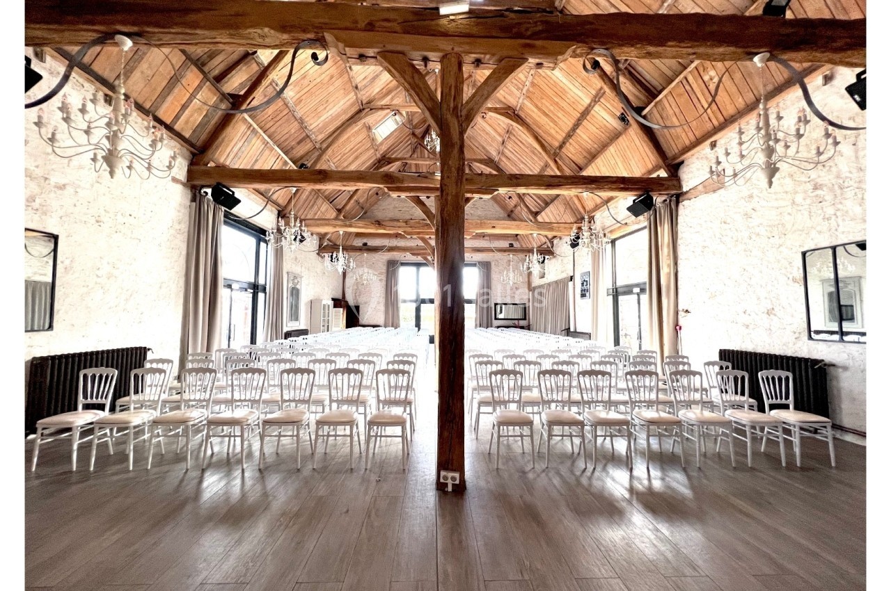 Salle lumineuse avec charpente en bois apparente, rangées de chaises blanches alignées pour un événement ou une réunion.