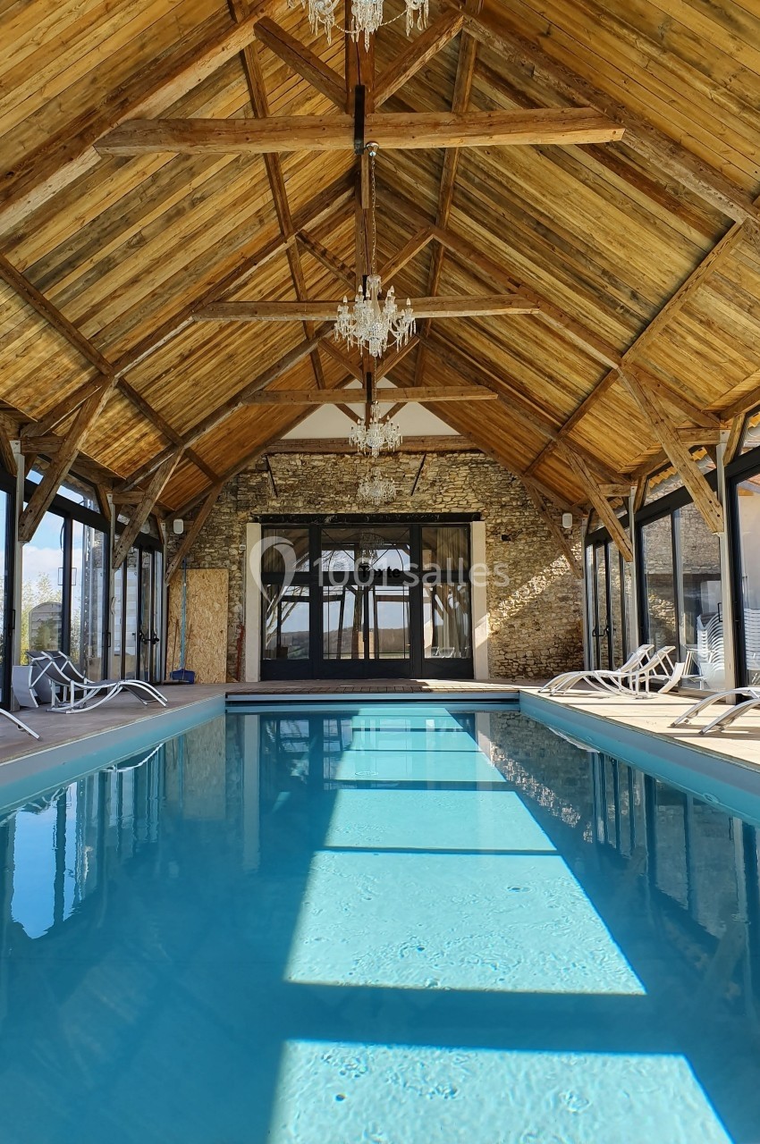 Piscine intérieure avec toit en bois, grandes baies vitrées et chaises longues disposées sur les côtés.