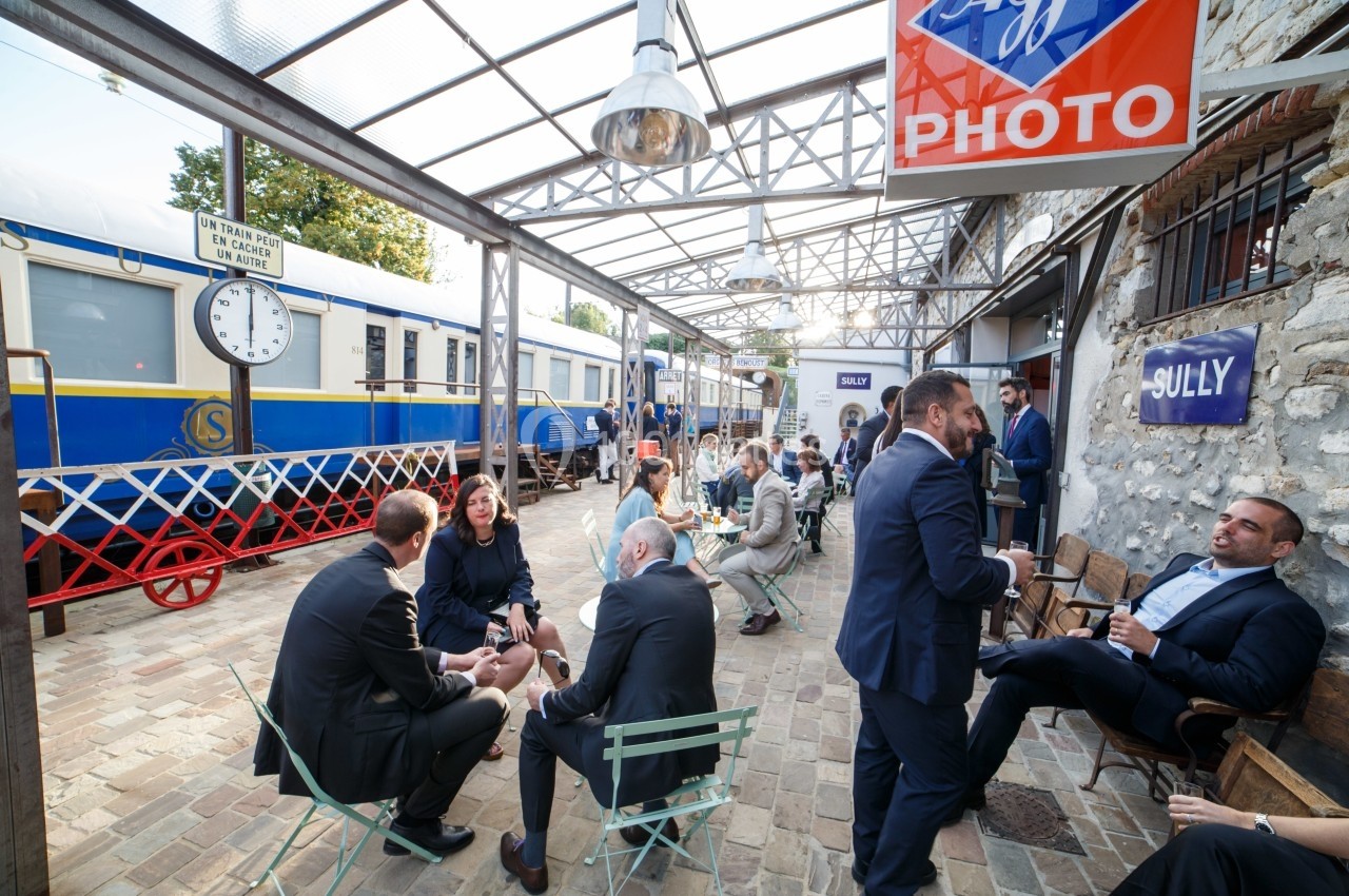 Personnes discutant en petits groupes sur une terrasse pavée près d'un train bleu et blanc dans une ambiance conviviale.