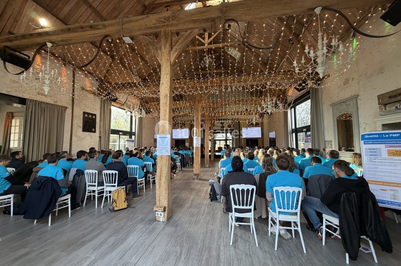 Salle en bois avec des participants assis en t-shirts bleus, assistant à une présentation projetée sur plusieurs écrans.