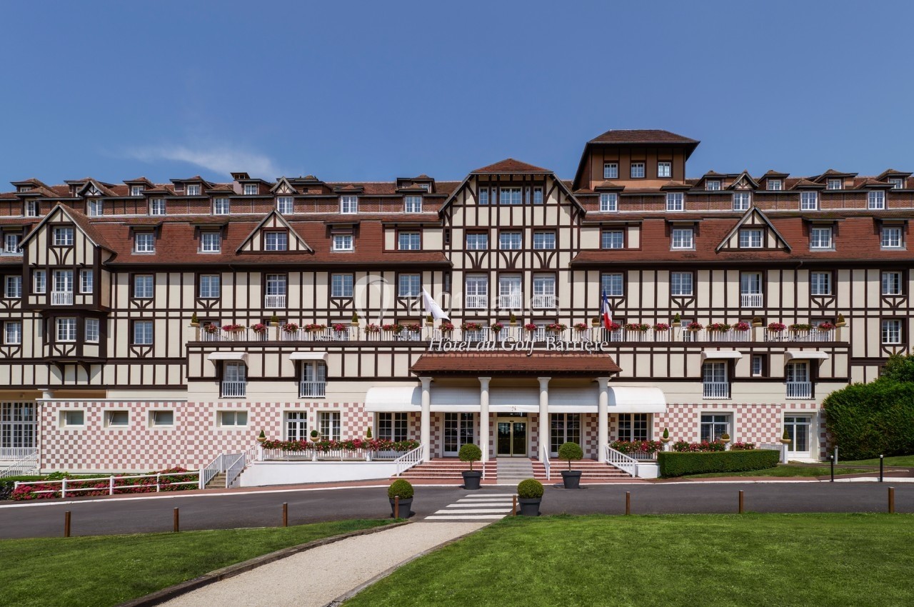 Façade d'un grand bâtiment à colombages avec balcons fleuris, entouré d'une pelouse et d'un chemin pavé.