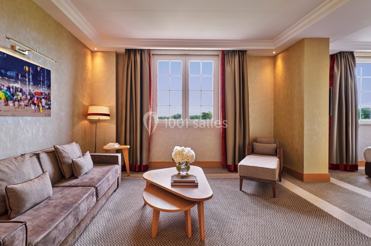 Salon lumineux avec canapé, fauteuil, table basse, lampadaire et fenêtres donnant sur un paysage verdoyant.