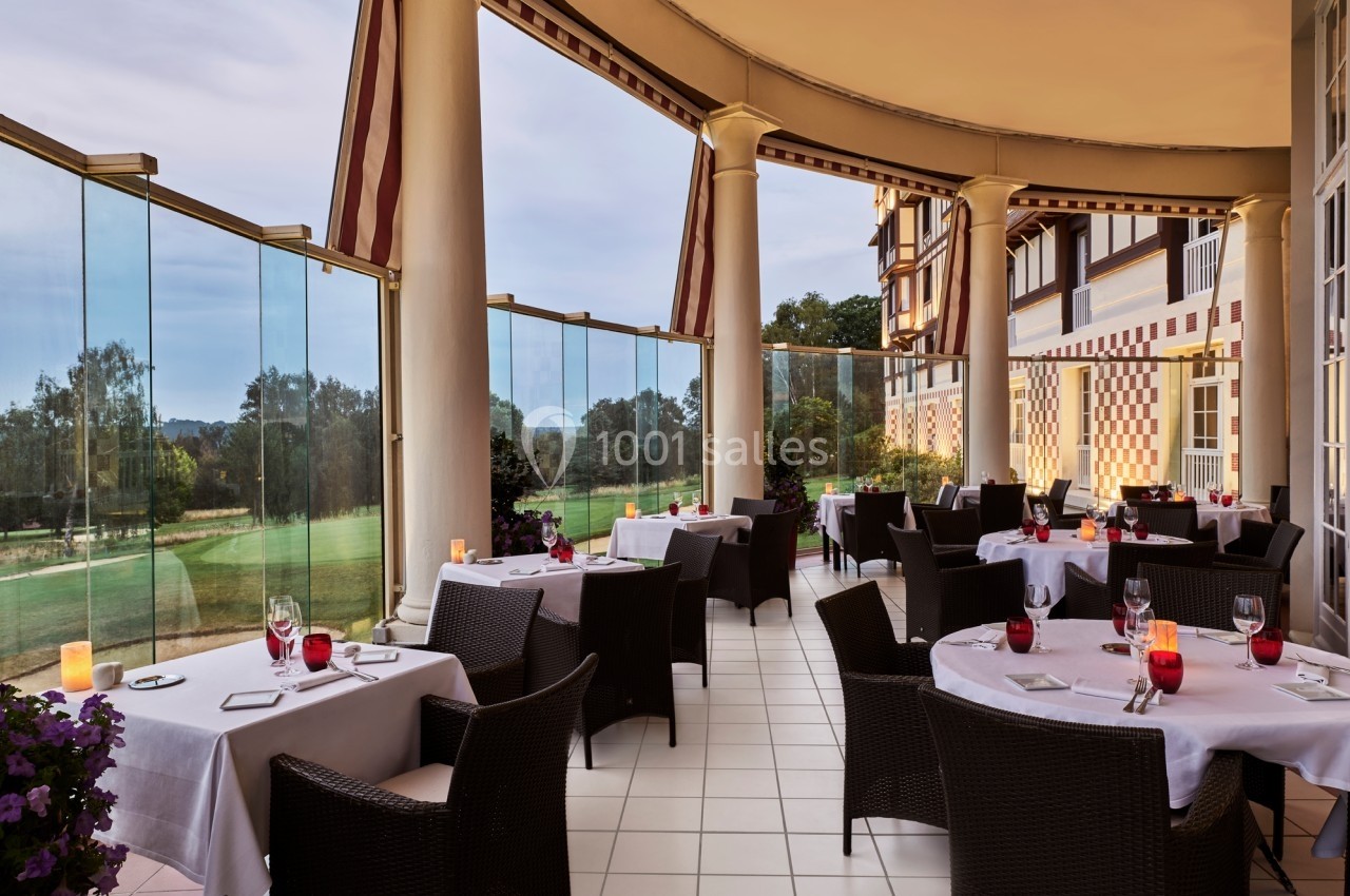Terrasse élégante avec tables dressées, chaises en osier, vue sur un jardin et balustrade en verre.