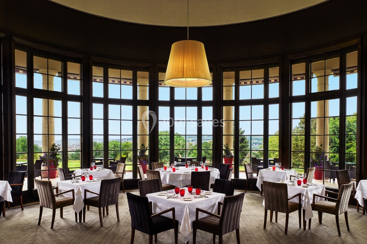 Salle de restaurant élégante avec grandes baies vitrées, tables dressées et vue sur un paysage verdoyant.