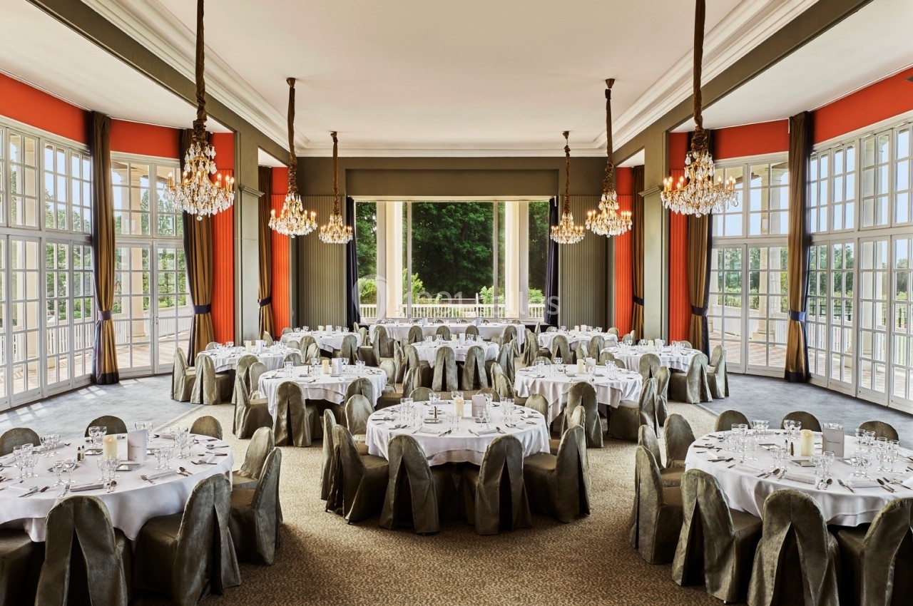 Salle de réception élégante avec tables rondes dressées, chaises couvertes et lustres, donnant sur un jardin verdoyant.