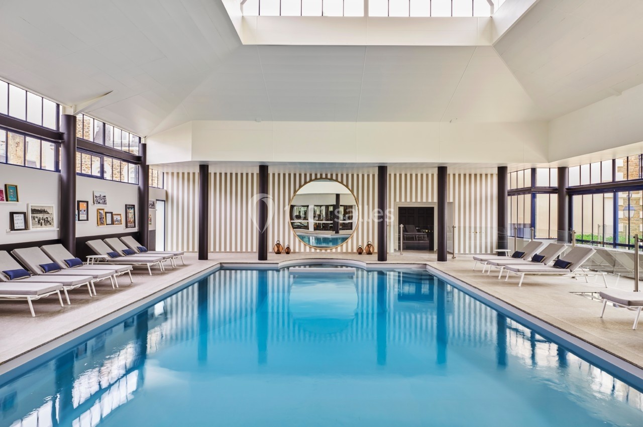Piscine intérieure lumineuse entourée de chaises longues, avec un grand miroir rond et des baies vitrées.