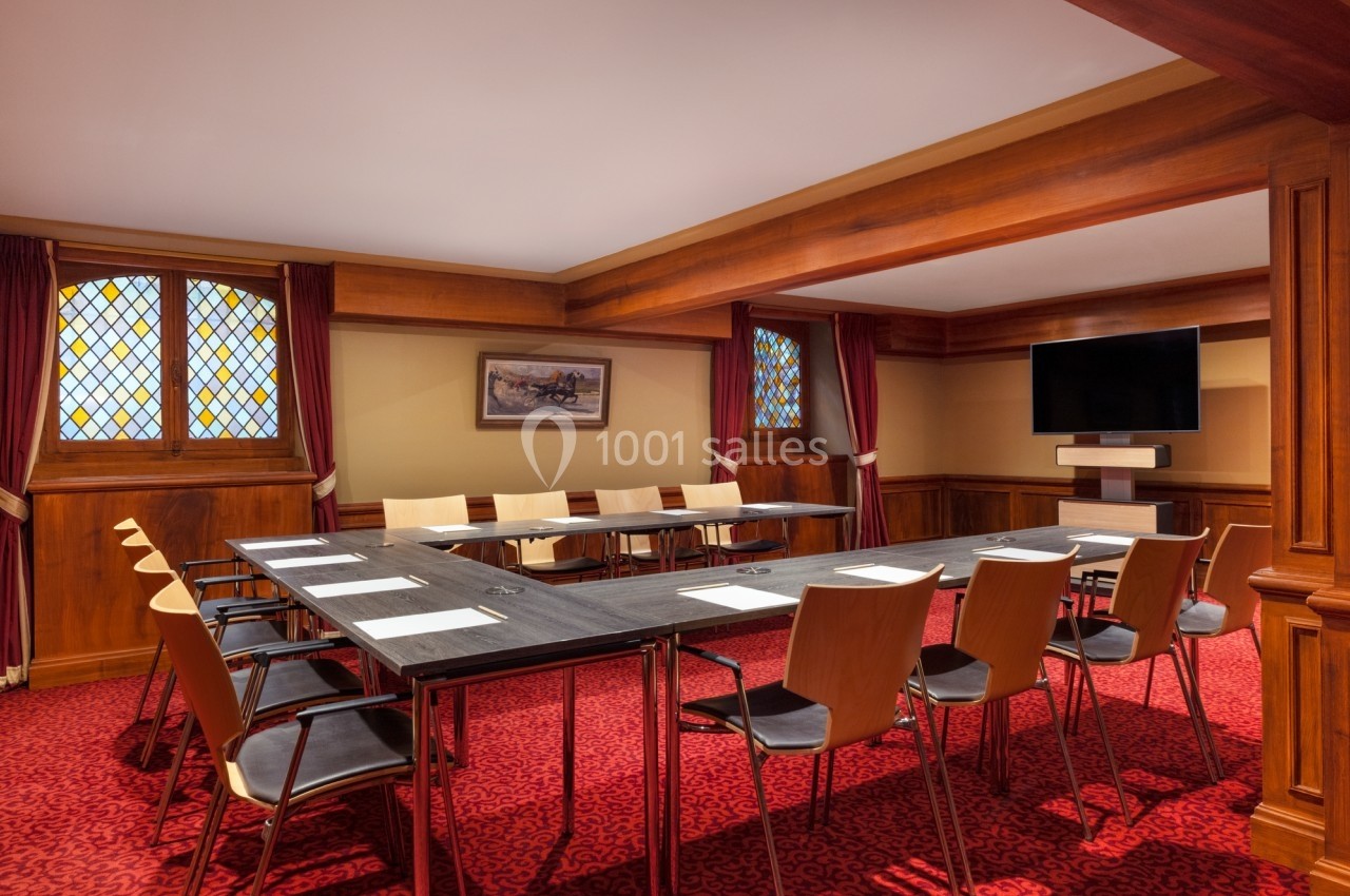 Salle de réunion avec table en U, chaises en bois, écran mural et fenêtres à vitraux, sur moquette rouge.