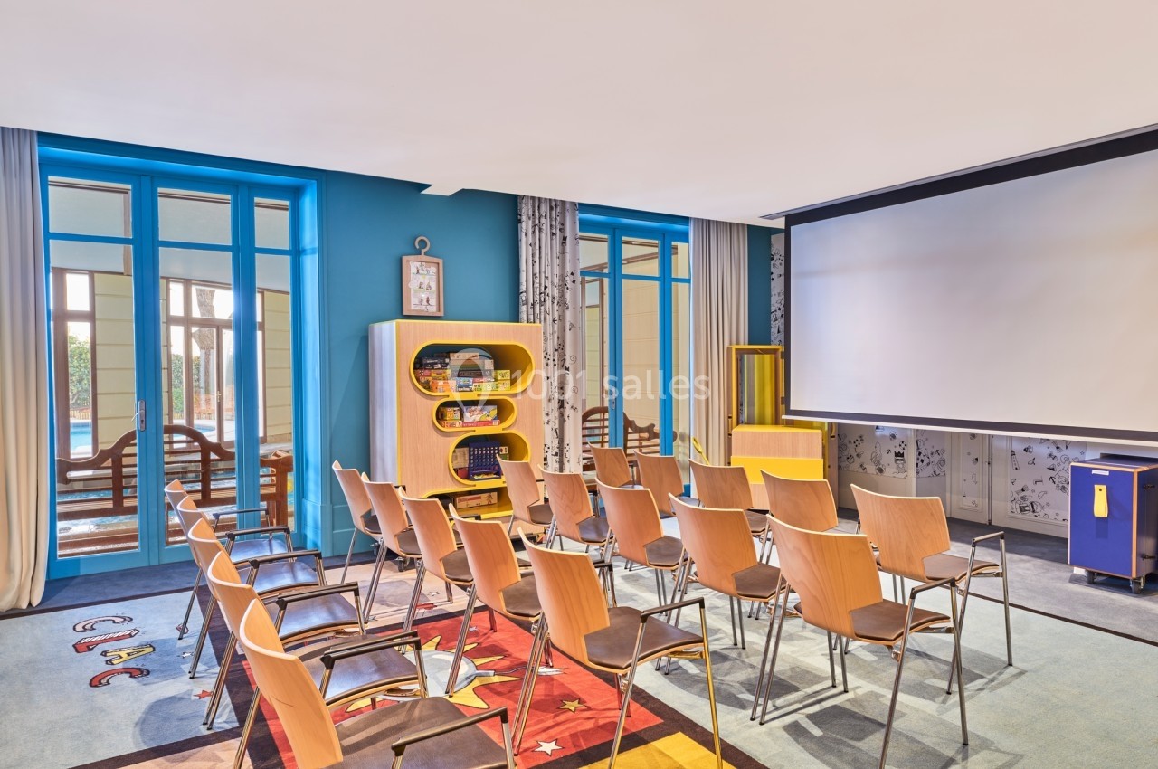 Salle de conférence lumineuse avec des chaises en bois, un écran de projection et des étagères contenant des jeux.