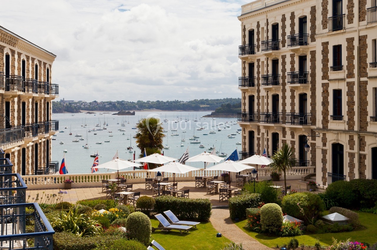 Vue sur une terrasse bordée de parasols et de jardins, donnant sur une baie avec des voiliers et des bâtiments en arrière…