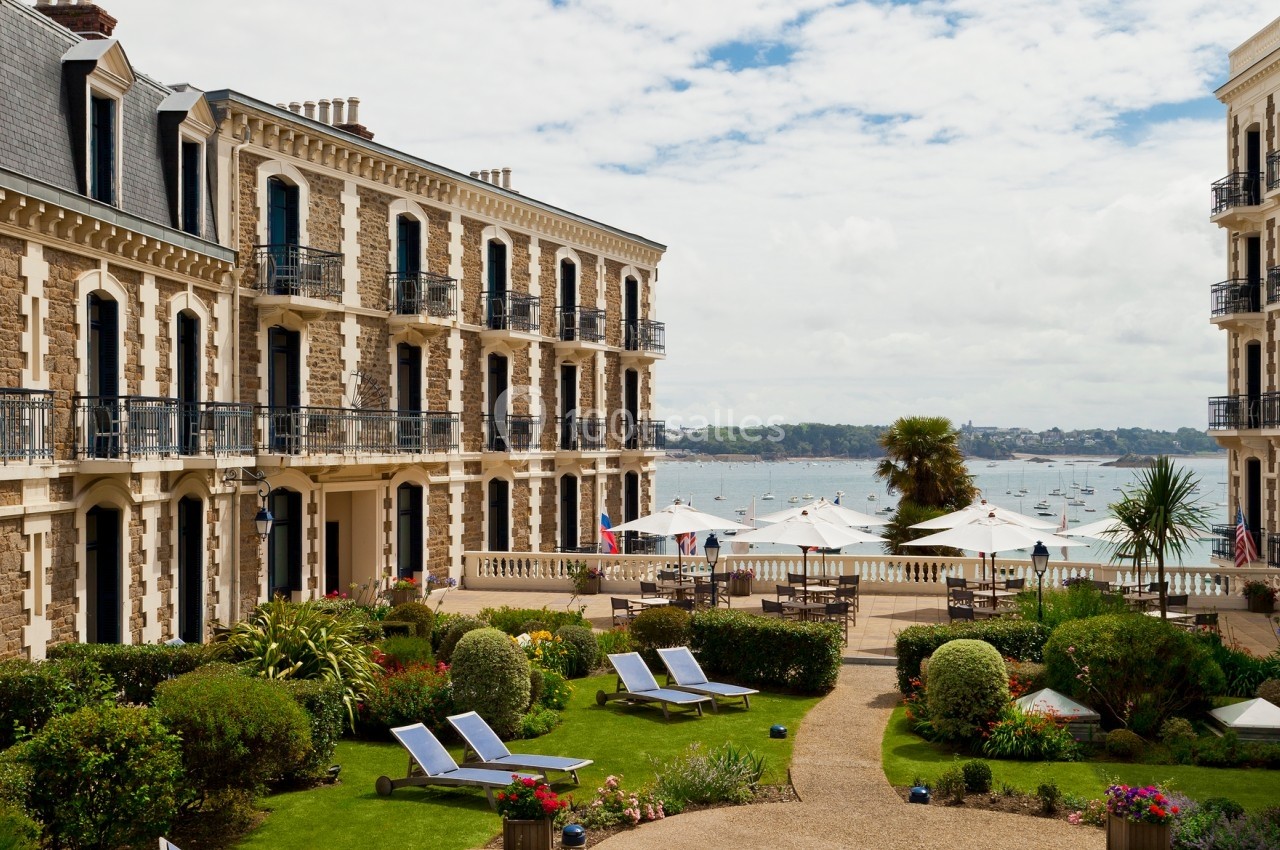 Cour intérieure d'un hôtel avec jardin aménagé, transats, parasols et vue sur la mer avec des voiliers au loin.