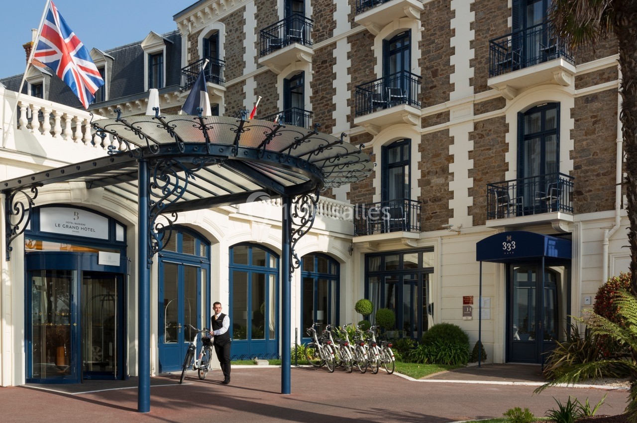 Entrée d'un hôtel de style classique avec un employé poussant un vélo près d'un porche en fer forgé.