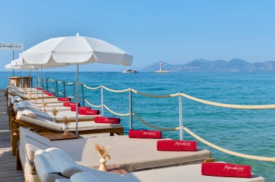 Transats avec coussins rouges alignés sur un ponton en bois, face à la mer, avec parasols blancs et vue sur un phare.