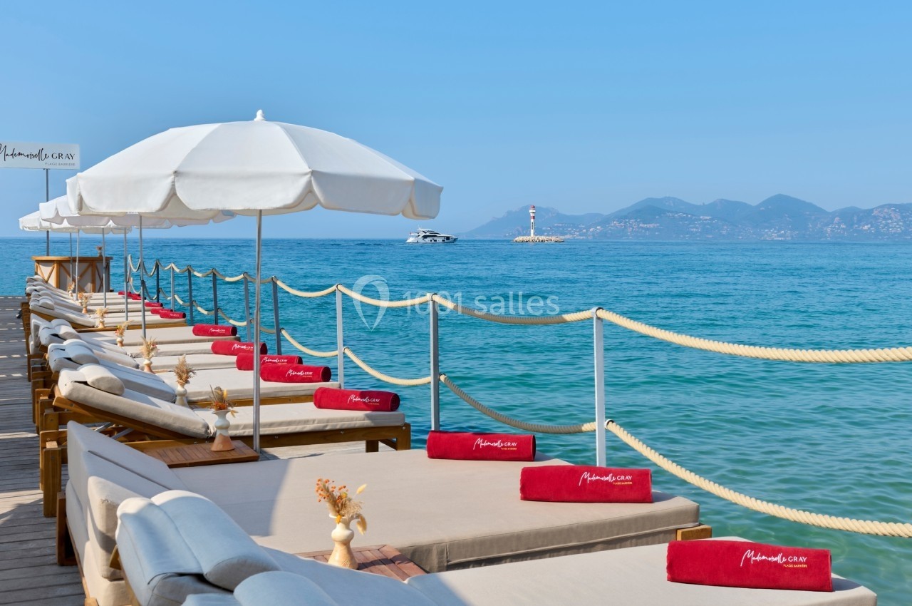 Transats avec coussins rouges alignés sur un ponton en bois, face à la mer, avec parasols blancs et vue sur un phare.