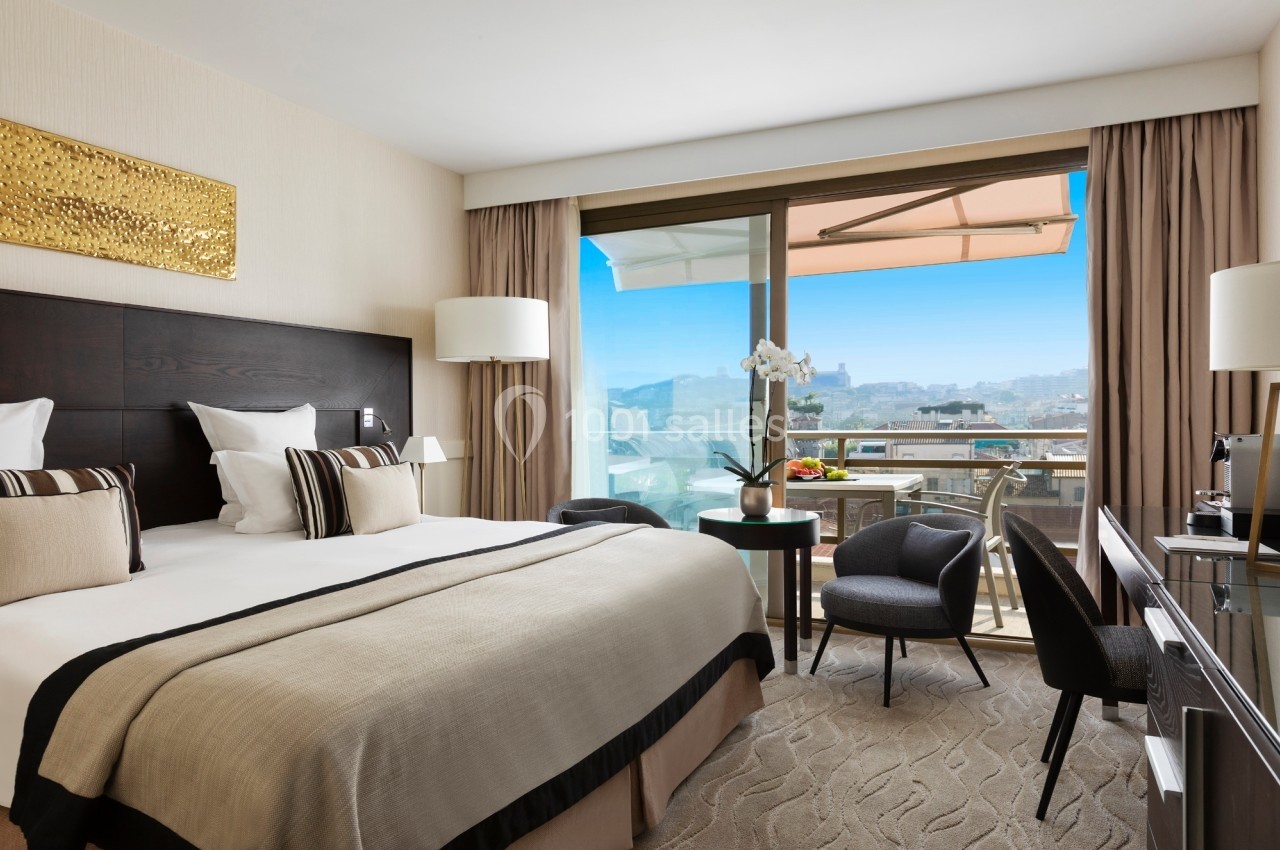 Chambre d'hôtel lumineuse avec lit double, balcon aménagé et vue sur la ville sous un ciel bleu.