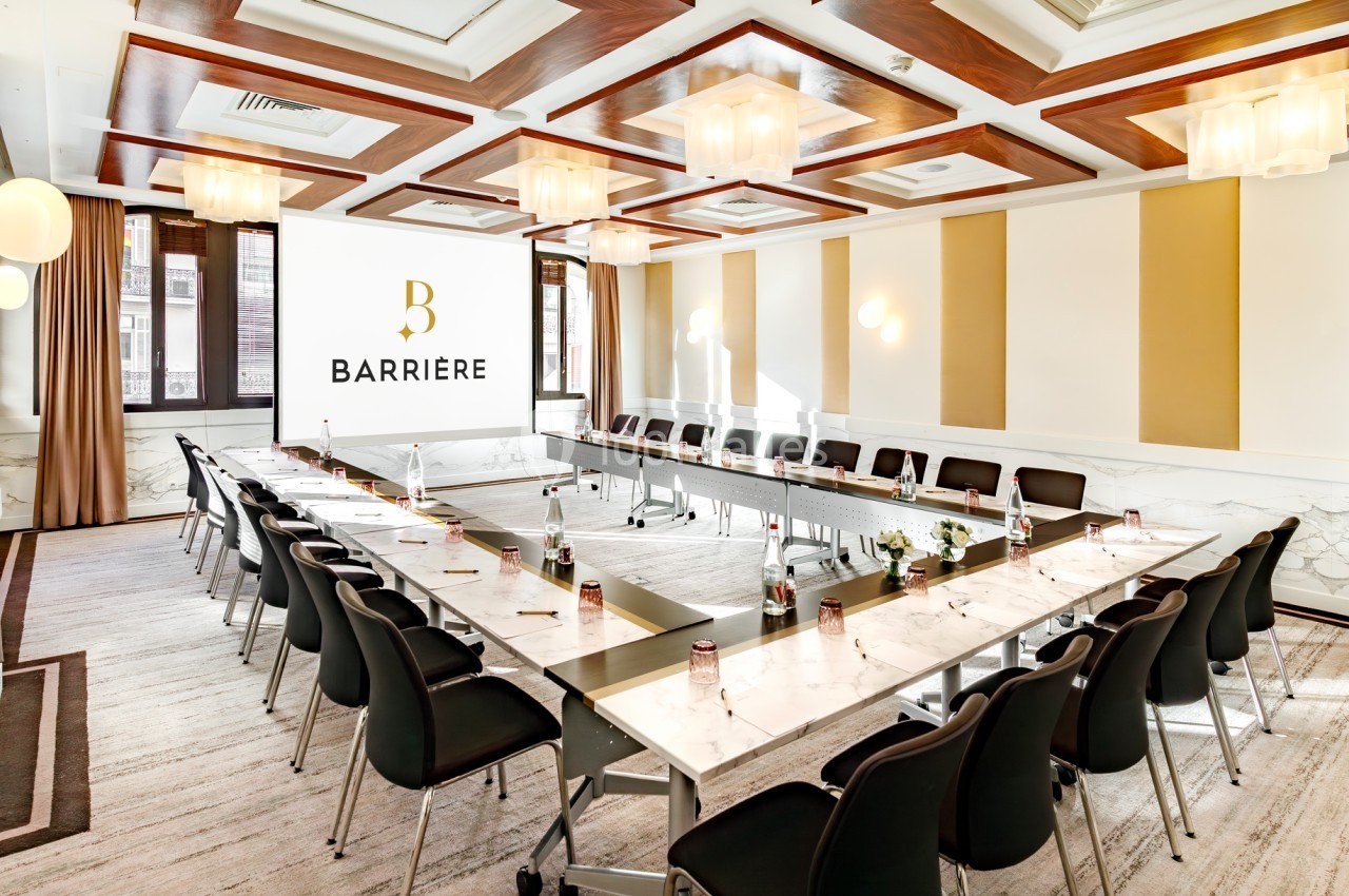 Salle de réunion lumineuse avec tables en U, chaises noires, écran de projection et éclairage moderne.