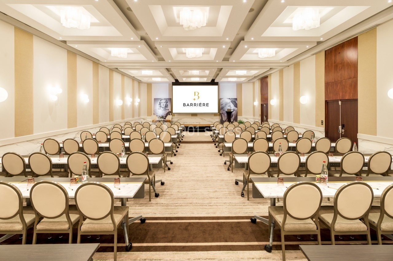 Salle de conférence lumineuse avec rangées de tables et chaises alignées face à un écran de présentation.