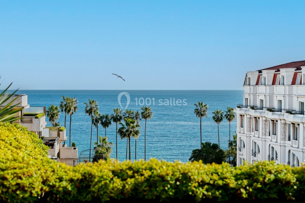 Vue sur la mer avec des palmiers, un bâtiment blanc et un ciel dégagé sous une lumière ensoleillée.