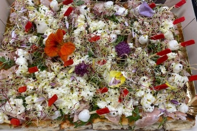Plateau garni de charcuteries, fromage frais, fleurs comestibles et pousses, disposés sur des tranches de pain.