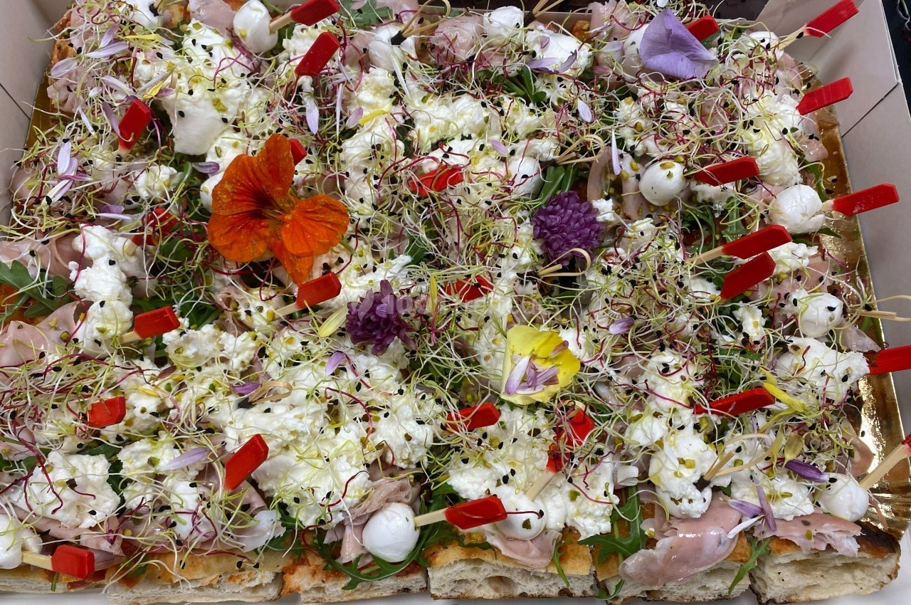 Pizza garnie de mozzarella, jambon, fleurs comestibles, pousses et brochettes rouges dans un plateau.