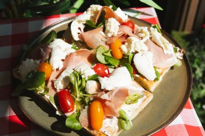 Assiette de tartines garnies de jambon cru, mozzarella, tomates cerises et mâche sur une nappe à carreaux rouges.
