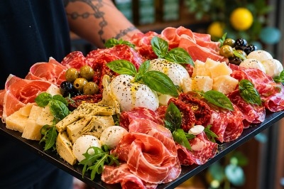 Plateau de charcuteries et fromages avec burrata, olives, tomates séchées, artichauts et basilic, tenu par une personne.