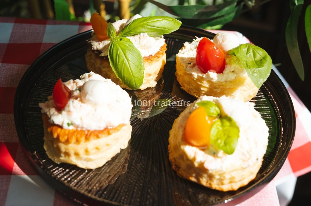 Assortiment de feuilletés garnis de fromage frais, tomates cerises et feuilles de basilic, sur une assiette noire.
