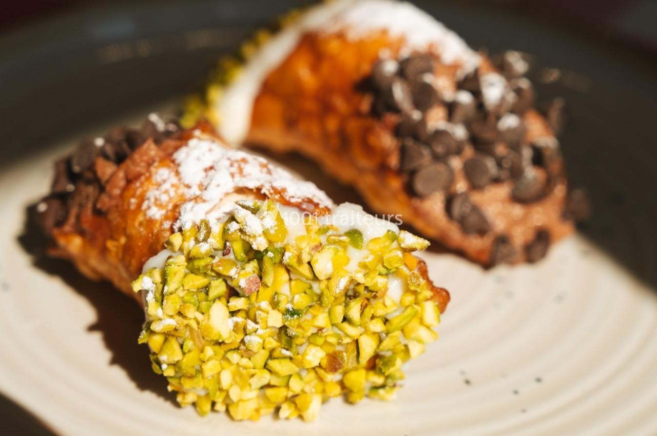 Deux cannoli garnis de crème, l'un décoré de pistaches hachées, l'autre de pépites de chocolat, sur une assiette.
