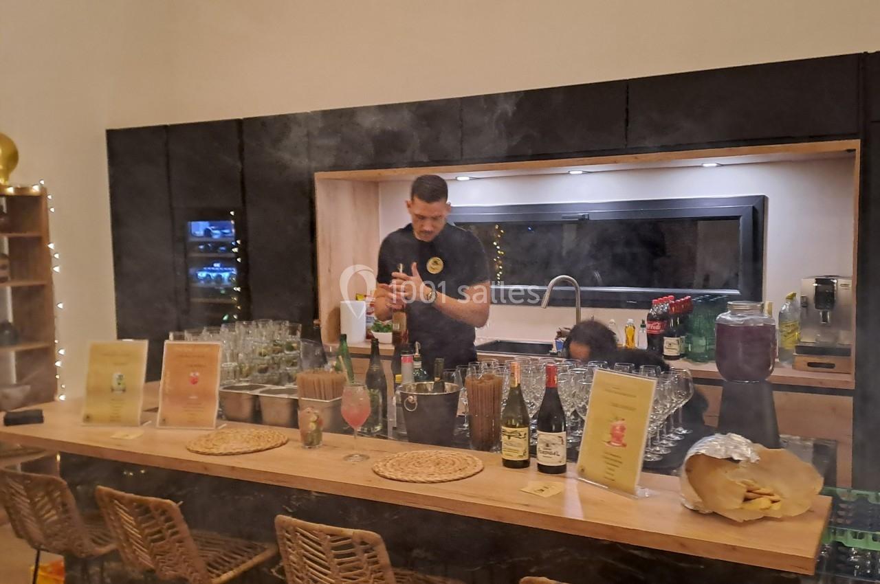 Un barman prépare des cocktails derrière un comptoir en bois, entouré de bouteilles, verres et menus.