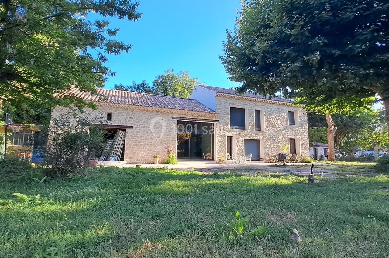 Maison en pierre avec toit en tuiles, grandes baies vitrées et jardin arboré sous un ciel dégagé.